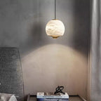 Minimalist alabaster mini pendant light with a gold frame and glowing LED, hanging above a bedside table.