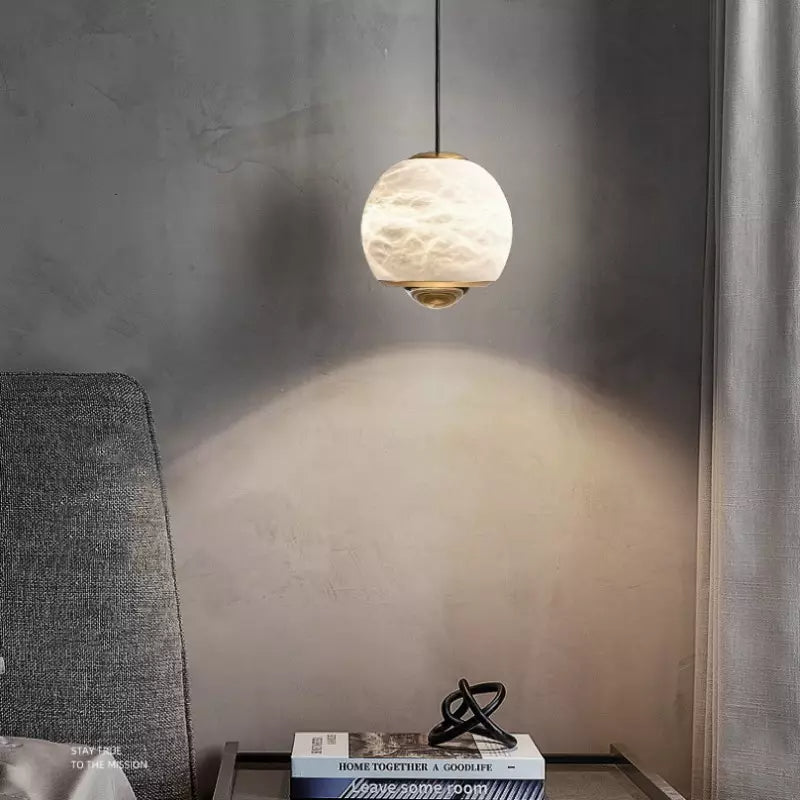 Minimalist alabaster mini pendant light with a gold frame and glowing LED, hanging above a bedside table.