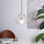 Sculptural clear glass pendant light with a rippled, tiered silhouette and warm filament bulb hanging over a dining table.