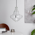Sculptural clear glass pendant light with a flowing, rippled silhouette and a central filament bulb hanging over a table.