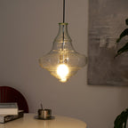 Sculptural clear glass pendant light with a rippled silhouette and warm glowing filament bulb hanging above a table.