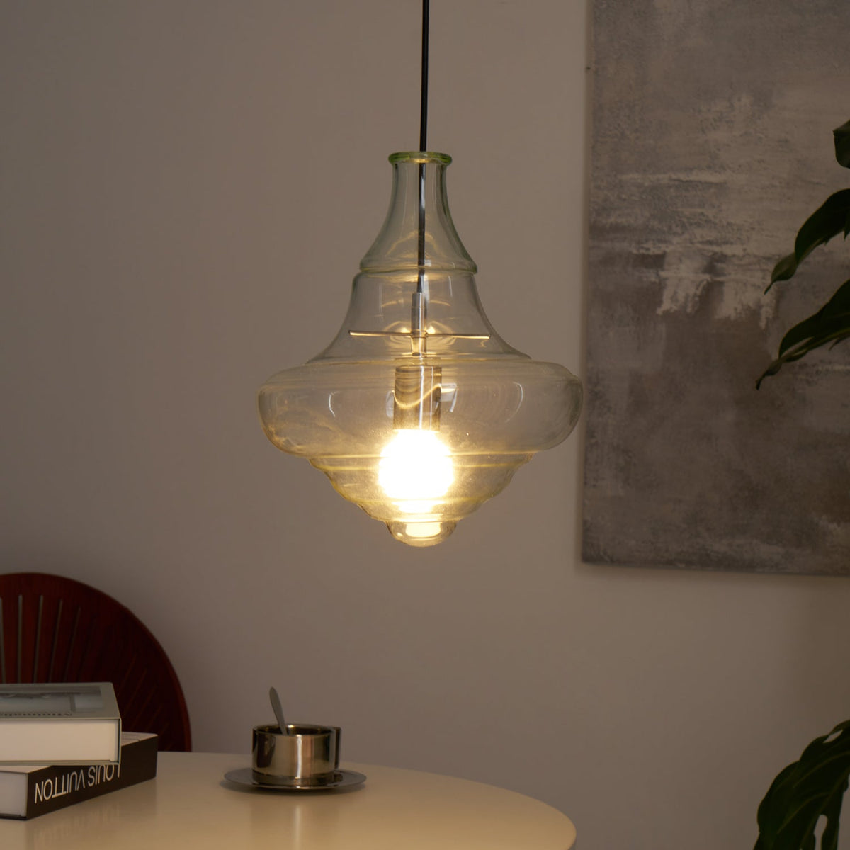Sculptural clear glass pendant light with a rippled silhouette and warm glowing filament bulb hanging above a table.