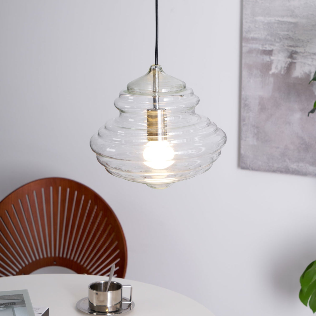 Sculptural clear glass pendant light with a rippled, tiered design and a warm glowing filament bulb hanging above a table.