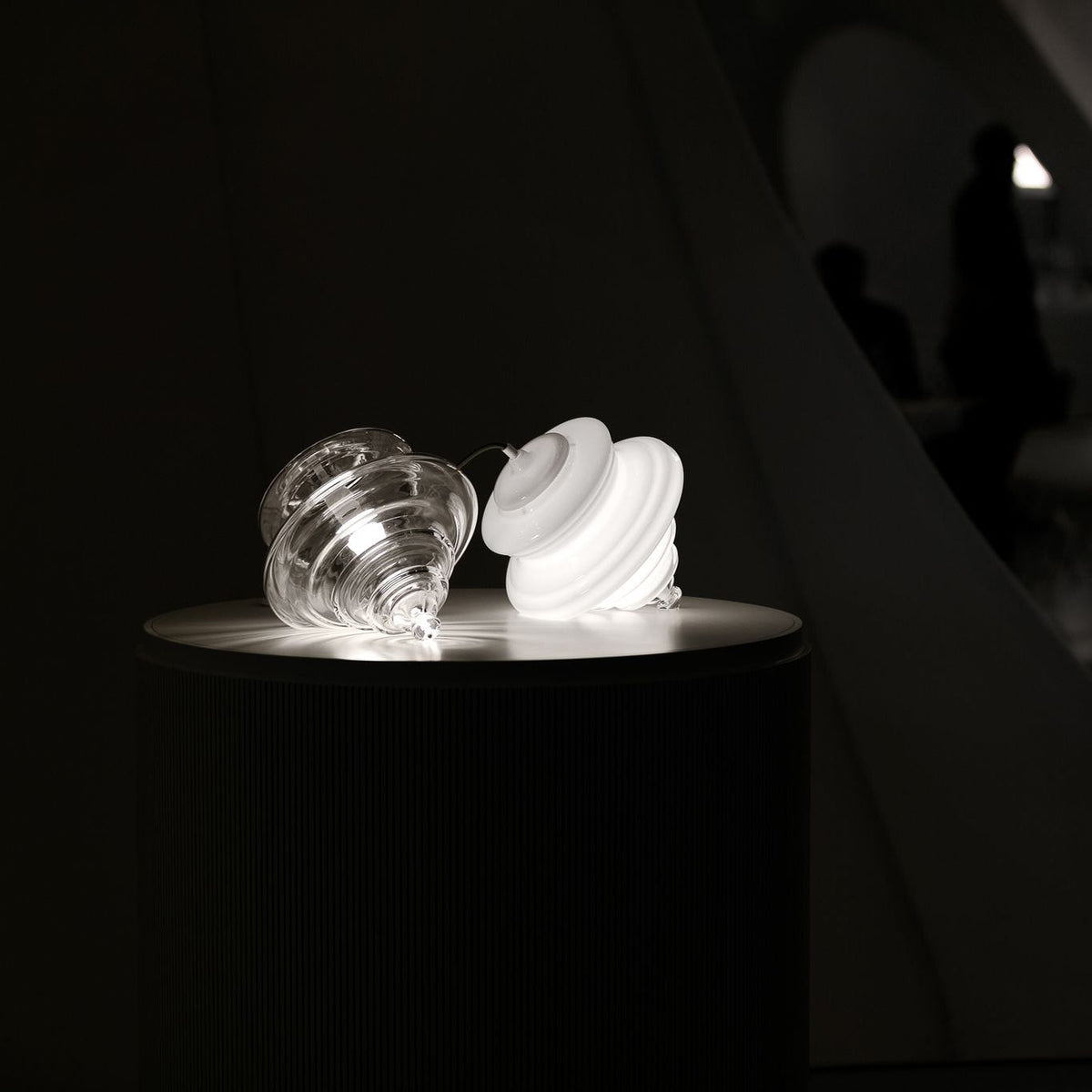 Two sculptural clear glass pendant lights with rippled, tiered silhouettes resting on a circular pedestal.