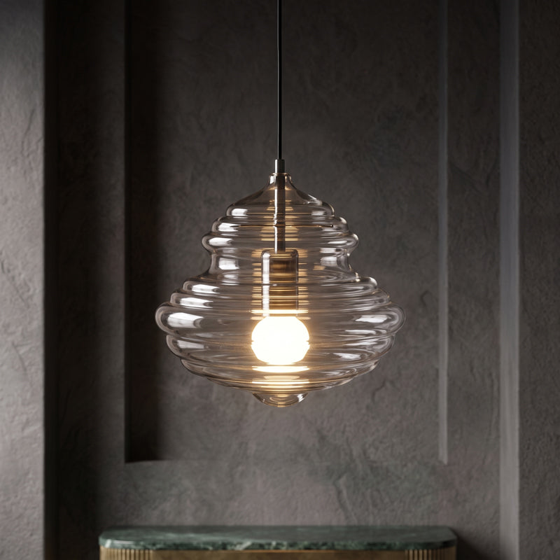 Sculptural clear glass pendant light with rippled texture and a warm glowing filament bulb against a dark wall.
