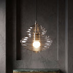 Sculptural clear glass pendant light with rippled texture and a warm glowing filament bulb against a dark wall.