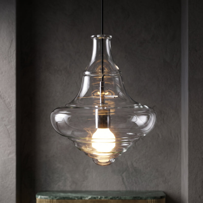 Sculptural clear glass pendant light with a rippled silhouette and warm filament bulb against a dark background.