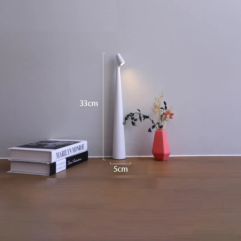 Slim white cordless LED table lamp, 33cm tall with a minimalist design, placed on a wooden surface next to books and a vase.