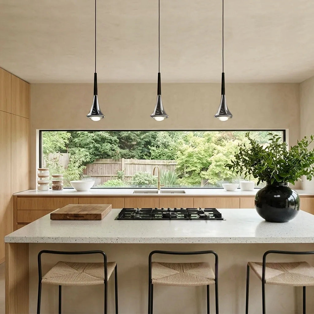 Three chrome teardrop LED pendant lights hanging over a modern kitchen island with a view of a garden.