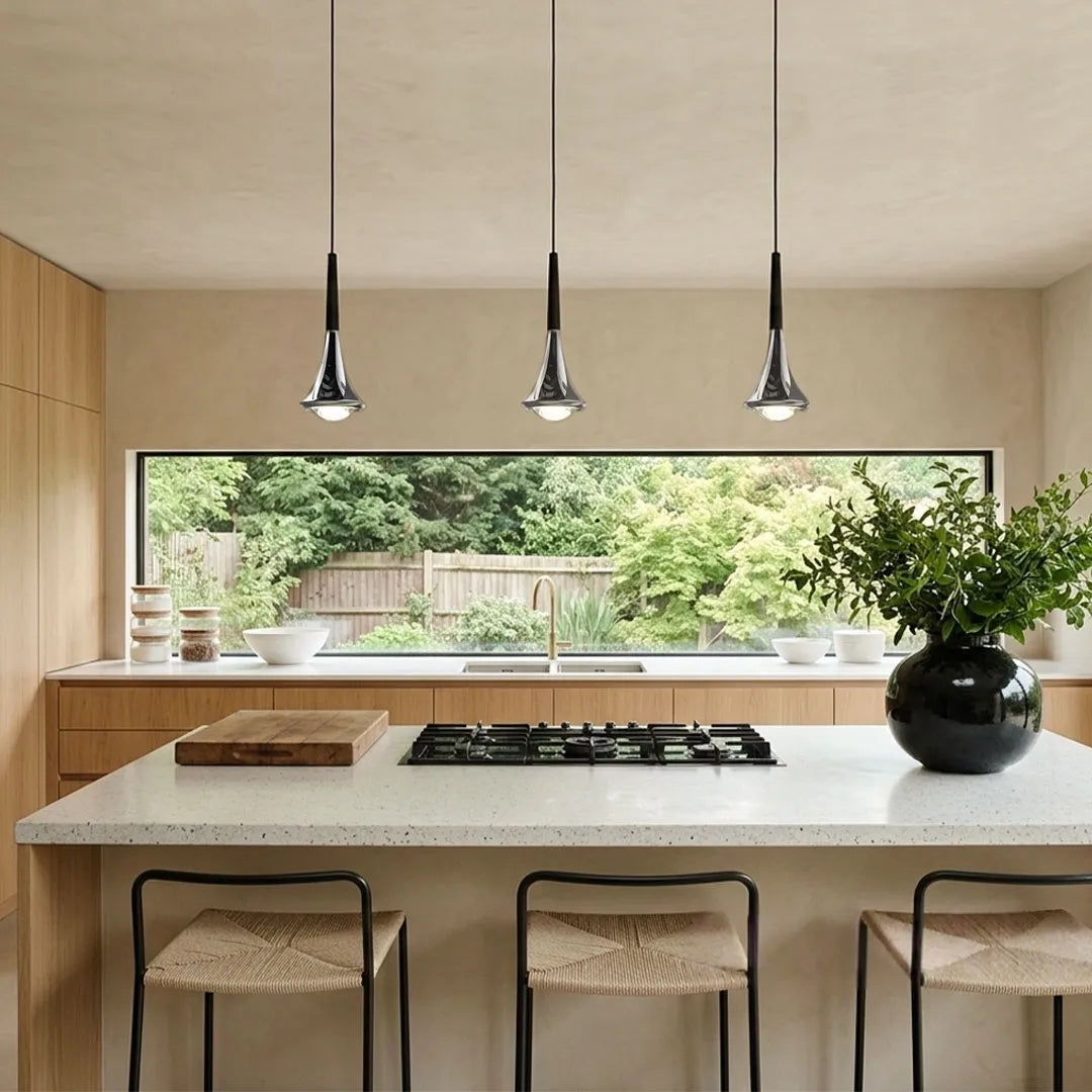 Three chrome teardrop LED pendant lights hanging over a modern kitchen island with a view of a garden.