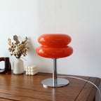 Retro orange glass table lamp with a rounded 70s space age design on a polished chrome base sitting on a wooden table.