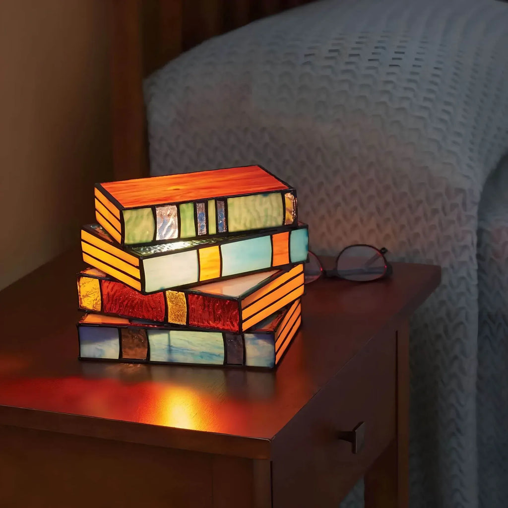 Vintage stained glass table lamp shaped like a stack of glowing, multicolored books on a wooden nightstand.