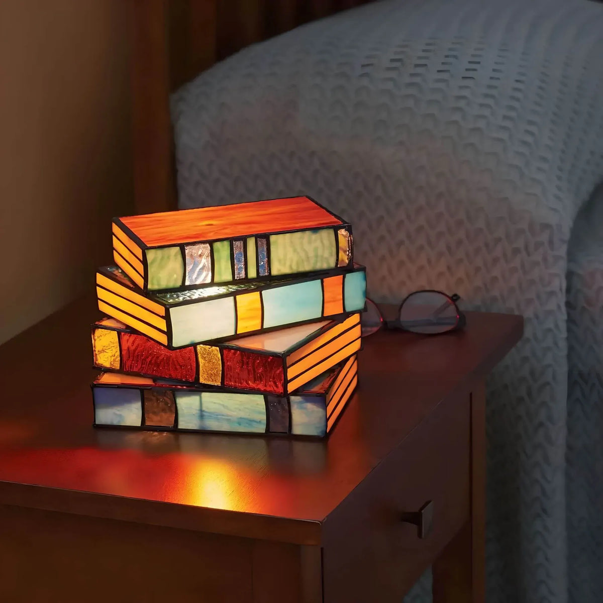 Vintage stained glass table lamp shaped like a stack of glowing, multicolored books on a wooden nightstand.
